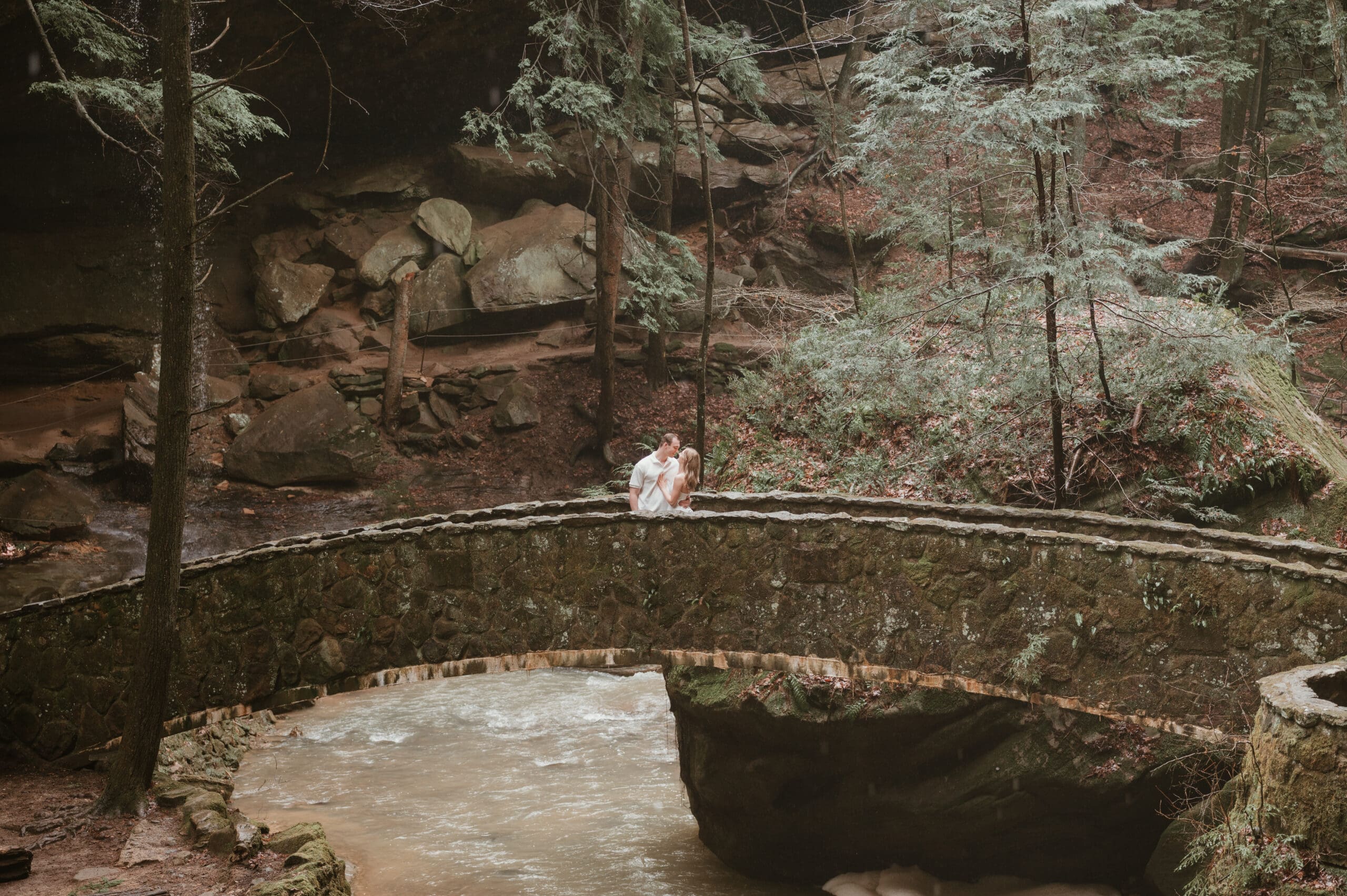 Couple kissing on the stone bridge at Old Man's Cave in Hocking Hills, Ohio during their elopement. Surrounded by evergreen trees, mossy rocks, and a flowing creek, this romantic moment captures the wild and intimate beauty of a forest elopement in Southern Ohio